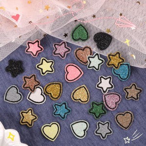 Könnte beinhalten: Eine Sammlung glitzernder Sterne und herzförmiger Verzierungen in verschiedenen Farben, darunter Rosa, Gold, Grün und Silber. Jede Form hat eine schwarze Umrandung. Die Formen sind auf einem blauen Stoff verstreut.