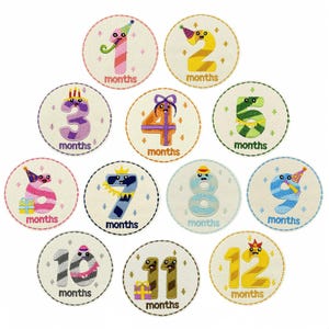 Pode incluir: Um conjunto de doze patches bordados, cada um com um número de um a doze com um rosto caprichoso e um chapéu de festa. A palavra "months" é bordada abaixo de cada número. Os patches são circulares com um fundo creme.