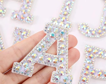 Può includere: Primo piano della lettera "A" ricoperta di strass iridescenti. La lettera è bianca con una consistenza simile al feltro ed è circondata da altre lettere con abbellimenti simili. Gli strass riflettono vari colori.