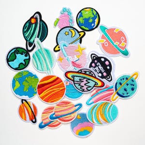 May include: A selection of embroidered planet patches in various colours. The patches feature planets with rings, Earth, and other celestial designs. The patches are outlined in black and have a white background.