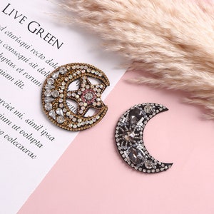 Può includere: Due toppe celesti decorative. Una è una luna crescente con una stella, impreziosita da strass dorati, trasparenti e rosa. L'altra è a forma di luna crescente con strass grigi e trasparenti. Il testo "LIVE GREEN" è visibile.