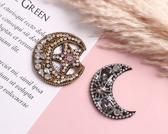 Puede incluir: Dos parches celestiales decorativos. Uno es una luna creciente con una estrella, adornada con pedrería dorada, transparente y rosa. El otro es una forma de media luna con pedrería gris y transparente. El texto "LIVE GREEN" es visible.