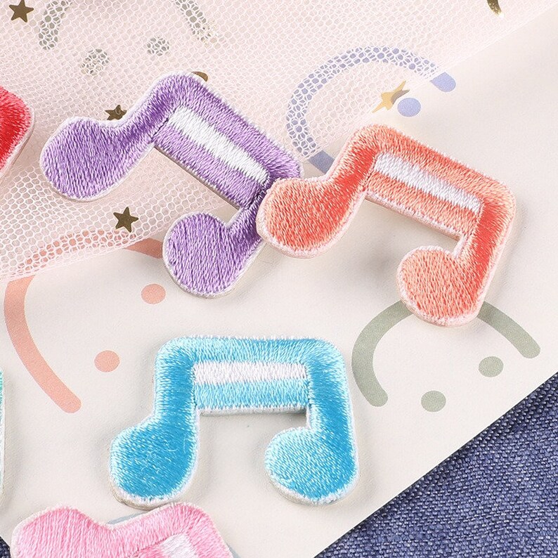 May include: Assortment of colourful embroidered iron-on patches shaped like musical notes. The patches are in shades of purple, pink, blue, and orange with white accents.