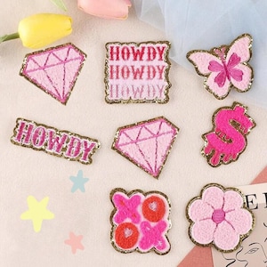 May include: Nine pink and gold glitter iron-on patches with various designs, including a diamond, a butterfly, a flower, a dollar sign, and the word "Howdy" repeated three times.