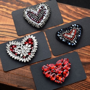 Può includere: Quattro decorazioni a forma di cuore in perline su cartoncino nero. Le decorazioni sono fatte di perline rosse, bianche e blu.