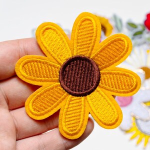 Op de afbeelding: Geborduurde gele zonnebloemplaats met een bruin centrum. De patch heeft een gedetailleerd gestikt ontwerp en een viltachtige textuur. De zonnebloem heeft acht bloemblaadjes en is waarschijnlijk voor decoratieve doeleinden.