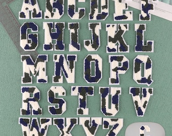 May include: A set of 26 white felt letters with a camouflage pattern in shades of green and blue. The letters are arranged in alphabetical order.
