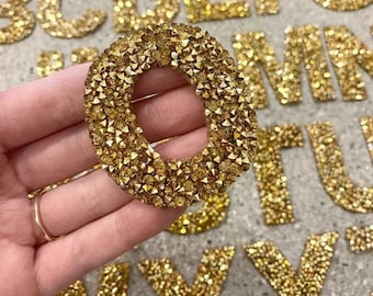 Può includere: Una lettera O dorata fatta di strass. La lettera è tenuta in una mano ed è circondata da altre lettere dell'alfabeto.