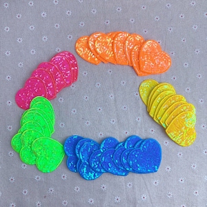 May include: Assortment of colorful heart-shaped sequins in various colors, including pink, orange, yellow, green, and blue. The sequins are arranged in a circle on a light gray fabric background.