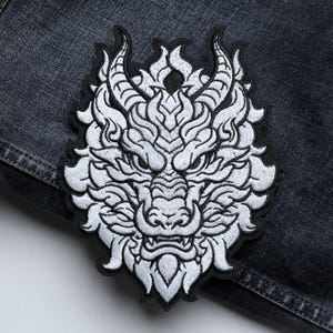 May include: Embroidered white dragon patch with intricate details, including horns, a snout, and sharp teeth. The patch is set against a dark denim background, showcasing the detailed stitching and design. The dragon's face is centered, with a black border.
