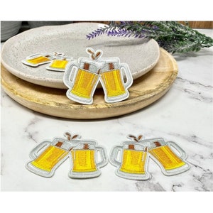 Op de afbeelding: Vier geborduurde strijkpatches met twee bierglazen die klinken. De patches zijn wit met geel bier en bruine accenten.