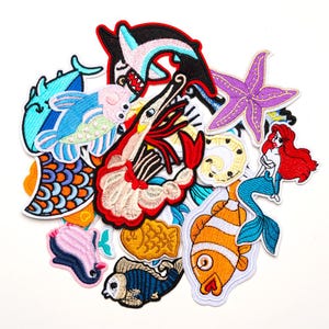 Op de afbeelding: Een verzameling geborduurde patches met verschillende zeedieren en personages. Ontwerpen omvatten een haai, garnaal, zeester, zeemeermin en verschillende soorten vissen in levendige kleuren zoals blauw, rood, oranje en paars. Ideaal voor het versieren van kleding of accessoires.