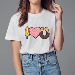 May include: White t-shirt with a graphic of a corn dog, a heart-shaped concha, and a churro spelling out "I love you".