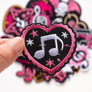 Può includere: Una toppa ricamata a forma di cuore con un centro nero che presenta una nota musicale viola chiaro e piccole stelle bianche. La toppa ha un bordo smerlato rosa. Altre toppe ricamate assortite sono visibili sullo sfondo.