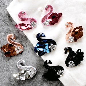 May include: Assortment of eight swan-shaped fabric patches with rhinestone embellishments. The patches are in various colors, including pink, blue, black, and gold.