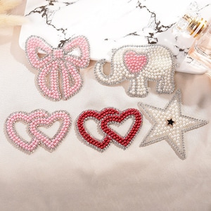 May include: Five pearl and rhinestone iron-on patches in various shapes: a bow, an elephant, two hearts, and a star. The patches are all pink, white, and red.