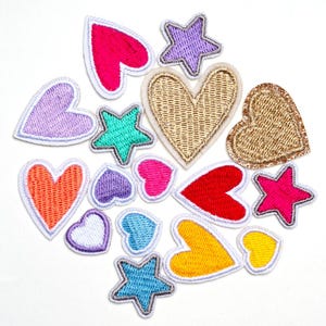 Op de afbeelding: Een verzameling kleurrijke geborduurde patches in verschillende vormen, waaronder harten en sterren. De patches hebben een verscheidenheid aan kleuren, zoals roze, rood, goud, paars, blauw en geel, met witte stiksels.
