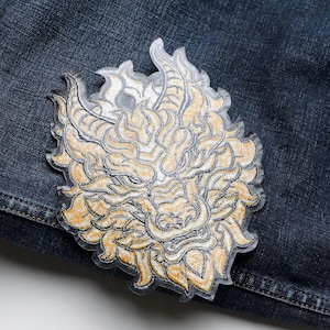 May include: Embroidered patch featuring a detailed dragon design in shades of gold and cream. The patch is placed on a dark blue denim background, showcasing the intricate stitching and design. The dragon's face is the focal point.