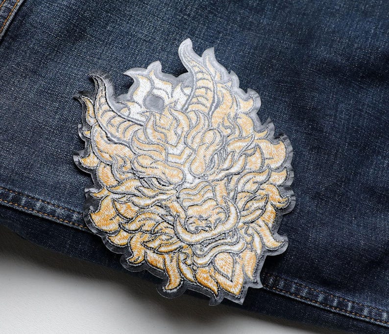 May include: Embroidered patch featuring a detailed dragon design in shades of gold and cream. The patch is placed on a dark blue denim background, showcasing the intricate stitching and design. The dragon's face is the focal point.