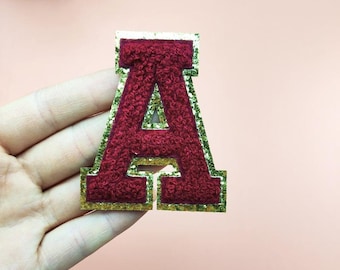 May include: A maroon letter 'A' patch with a gold glitter border. The letter has a textured, fuzzy appearance. The patch is a decorative item, likely for clothing or accessories.