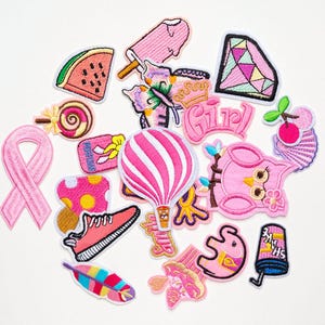 May include: A selection of pink embroidered patches featuring diverse designs such as a watermelon slice, ice lolly, butterfly, diamond, hot air balloon, and the word "Girl". Other patches include a trainer, feather, and a pink ribbon.