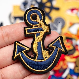 May include: Navy blue and gold embroidered anchor patch with a black border. The nautical-themed patch features detailed stitching, likely for application on clothing or accessories. Other patches are visible in the background.