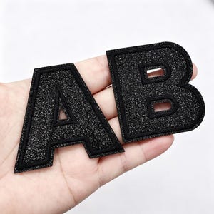 May include: Two black iron-on patches in the shape of the letters A and B. Each letter features a glittery, textured surface and is outlined with a thick, black embroidered border. The letters are presented as individual fabric appliqués.