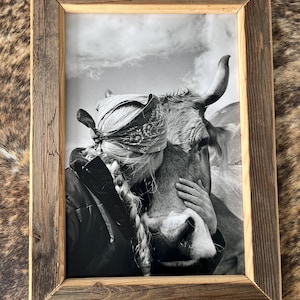 Könnte beinhalten: Schwarz-weiß gerahmte Fotografie einer Person, die eine Kuh umarmt. Das Foto befindet sich in einem rustikalen Holzrahmen. Die Kuh hat große Hörner und ist teilweise sichtbar. Die Person trägt ein Bandana und hat lange geflochtene Haare.