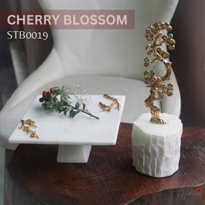 May include: A gold metal cherry blossom tree sculpture on a white marble base. The sculpture is on a wooden table with a white marble cake stand with gold metal branches on it. The text "CHERRY BLOSSOM" and "STB0019" is visible in the image.
