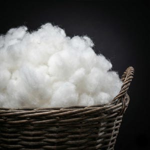 May include: A traditional wicker basket, brimming with soft, white cotton wool. The basket is a natural brown hue, and the cotton has a fluffy, cloud-like texture. The backdrop is a deep black.