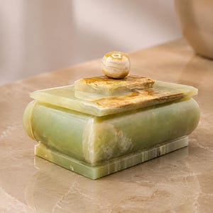 May include: A rectangular light green onyx stone box with a smooth, polished finish. The lid features a central square platform topped with a small, spherical knob. The stone displays natural translucent banding and warm brown veining throughout.