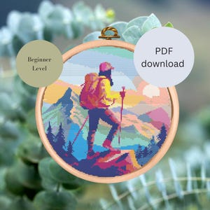 Könnte beinhalten: Ein Kreuzstichmuster mit einem Wanderer mit Rucksack, der auf einem Berggipfel steht, mit einem Sonnenuntergang im Hintergrund. Das Muster ist mit "Anfänger-Level" gekennzeichnet und als PDF-Download verfügbar.