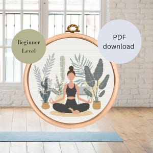 Könnte beinhalten: Ein Kreuzstichmuster mit einer Person in einer Yoga-Pose, umgeben von Pflanzen. Das Muster befindet sich in einem hölzernen Stickrahmen und ist mit "Anfänger-Level" beschriftet.