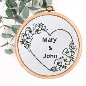 Könnte beinhalten: Schwarzer und weißer Stickrahmen mit einem Herzdesign und floralen Akzenten. Das Herz enthält die Namen "Mary & John".