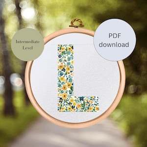 May include: A cross-stitch pattern for the letter L, featuring a floral design with yellow, blue, and green flowers. The pattern is displayed in a wooden embroidery hoop. The text "Intermediate Level" and "PDF download" are also visible.