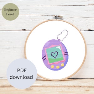 Könnte beinhalten: Ein Kreuzstichmuster eines lila und rosa Tamagotchis mit einem Herz auf dem Bildschirm. Das Muster ist in einem hölzernen Stickrahmen gerahmt. Der Text "PDF-Download" befindet sich auf einem hellblauen Kreis unter dem Rahmen.