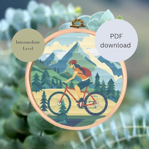 Könnte beinhalten: Kreuzstichmuster mit einer Frau, die mit dem Mountainbike in einem grünen Wald mit Bergen im Hintergrund fährt. Das Muster ist mit "Mittleres Niveau" gekennzeichnet und beinhaltet einen PDF-Download.