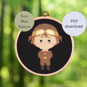 Könnte beinhalten: Ein Kreuzstichmuster eines Cartoon-Kindes, das eine braune Pilotenuniform und eine Schutzbrille trägt. Das Muster befindet sich in einem schwarzen Stickrahmen mit einem Holzrahmen. Der Text "Easy Mini Pattern" befindet sich auf einem grünen Kreis und der Text "PDF download" befindet sich auf einem blauen Kreis.