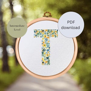 May include: A floral embroidery pattern for cross stitch of the letter T. The pattern is in a hoop and features yellow and blue flowers on a white background. The pattern is labeled as an intermediate level.