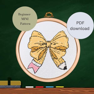 Könnte beinhalten: Kreuzstichmuster eines gelben Bogens und Bleistifts, in einem hölzernen Stickrahmen. Das Design beinhaltet einen rosa Radiergummi und den Text "Beginner MINI Pattern" und "PDF download".