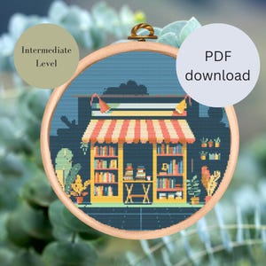 Könnte beinhalten: Ein Kreuzstichmuster mit einer farbenfrohen Illustration eines Buchladens mit Bücherregalen, einem Schild und Pflanzen. Das Muster ist mit "Mittleres Niveau" gekennzeichnet und als PDF-Download verfügbar.