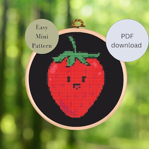 May include: A black and white cross stitch pattern of a red strawberry with a green stem and leaves. The strawberry has a cute cartoon face with a small smile and a tongue sticking out. The pattern is framed in a wooden hoop and the text "Easy Mini Pattern" and "PDF download" are visible in the image.