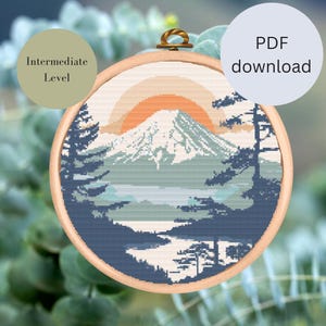 Könnte beinhalten: Ein Kreuzstich-Landschaftsdesign mit einem Berg, Bäumen und einem Sonnenuntergang in einem hölzernen Stickrahmen. Das Design enthält den Text "Intermediate Level" und "PDF download". Die Farbpalette umfasst Blau-, Grün-, Orange- und Cremetöne.