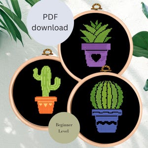 Könnte beinhalten: Drei schwarze Stickrahmen mit verschiedenen Kaktus-Designs. Die Rahmen befinden sich auf einem weißen Hintergrund. Die Designs beinhalten einen Kaktus mit einem herzförmigen Topf, einen Kaktus mit einem runden Topf und einen Kaktus mit einem hohen, dünnen Topf. Der Text "Anfänger-Level" befindet sich auf einem grünen Kreis unter den Rahmen.