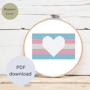 May include: A cross-stitch pattern featuring the transgender pride flag with a white heart in the center. The pattern is displayed in a wooden embroidery hoop on a white wood background. The text "Beginner Level" and "PDF download" are also visible.