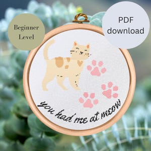 Könnte beinhalten: Ein Kreuzstichmuster mit einer Comic-Katze und dem Text "You had me at meow". Das Muster ist in einem Holzrahmen eingefasst und als Anfängerprojekt gekennzeichnet.