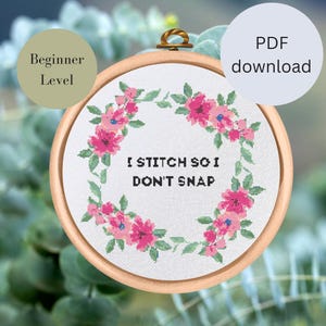 Könnte beinhalten: Ein weißer Stickrahmen mit einem rosafarbenen Blumenkranz und dem Text "Ich sticke, damit ich nicht ausraste".