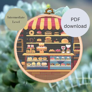 Könnte beinhalten: Ein Pixel-Art-Kreuzstichmuster einer Bäckerei mit einer rot-weiß gestreiften Markise. Das Schaufenster der Bäckerei zeigt eine Auswahl an Gebäck, Kuchen und Keksen. Das Muster ist mit "Mittleres Niveau" gekennzeichnet und als PDF-Download verfügbar.