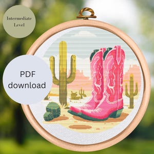 Könnte beinhalten: Ein Kreuzstich-Design in einem Holzrahmen zeigt rosa Cowboy-Stiefel in einer Wüstenlandschaft mit Kakteen. Der Text "PDF download" und "Intermediate Level" sind ebenfalls enthalten.