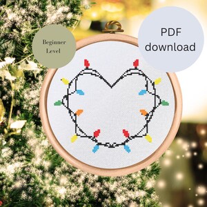 May include: A cross-stitch pattern of a heart-shaped string of Christmas lights in a wooden embroidery hoop. The pattern is labeled "Beginner Level" and is available as a PDF download.